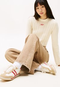 Jeune femme assise en tailleur, portant un pull crème en tricot torsadé, un pantalon beige à large jambe et des baskets blanc-beige avec des rayures rouges.