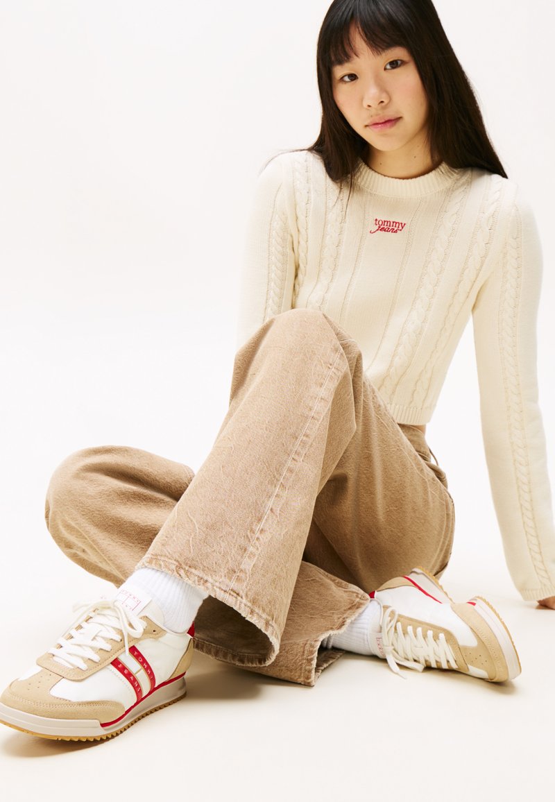 Jeune femme assise en tailleur, portant un pull crème en tricot torsadé, un pantalon beige à large jambe et des baskets blanc-beige avec des rayures rouges.