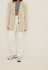 Beige oversized blazer met twee knopen, gestreept cropped topje, witte wijde broekspijpen en zilveren hardloopschoenen met zwarte accenten.