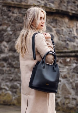 Black leather handbag with dual handles, gold logo detail, and smooth texture. Styled with a beige coat against a stone wall backdrop.