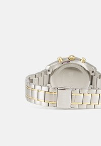 Silberne Edelstahl-Uhr mit einem zwei-farbigen Armband, das goldene Akzente aufweist, runder Gehäuserand und drei Druckknöpfe oben.