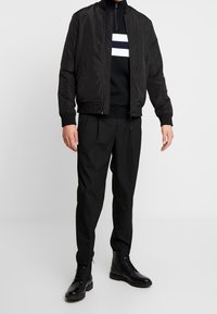 Blouson aviateur noir à finition lisse, porté sur un pull noir avec des rayures horizontales blanches et bleu marine, associé à un pantalon noir et des bottes.