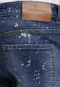 Jeans av denim med en urtvättad blå nyans, med vita sprutmönster, en brun läderlapp vid midjan och orangea sömnadsdetaljer.