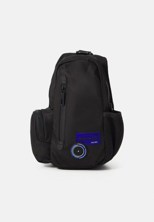 Black backpack made of durable material, featuring zippers, mesh side pockets, and a blue logo patch with circular detailing. Compact design.