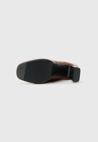 Zapato de tacón de cuero marrón tumbado de lado, mostrando una suela texturizada negra y un tacón cuadrado sobre un fondo claro.