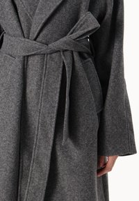 Siva volnena obleka z mehkobo, z pasom, širokimi rokavi in sprednjimi žepi. Ima šalikov ovratnik in minimalne detail šivanja.