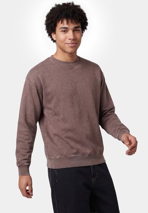 Junger Mann mit lockigem Haar, der einen braunen Pullover mit langen Ärmeln und schwarze Hosen trägt, steht vor einem einfarbigen weißen Hintergrund.