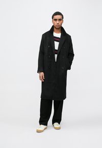 Černý dvouřadý kabát z tkaniny s příměsí vlny, s širokým klopovým límcem a rovnou siluetou, nošený přes pruhovaný svetr a černé kalhoty.