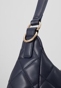 Marinblå läderväska med quiltat mönster, utrustad med en guldmetallring och matchande detalj på banden. Slät textur med kontrasterande stygn.