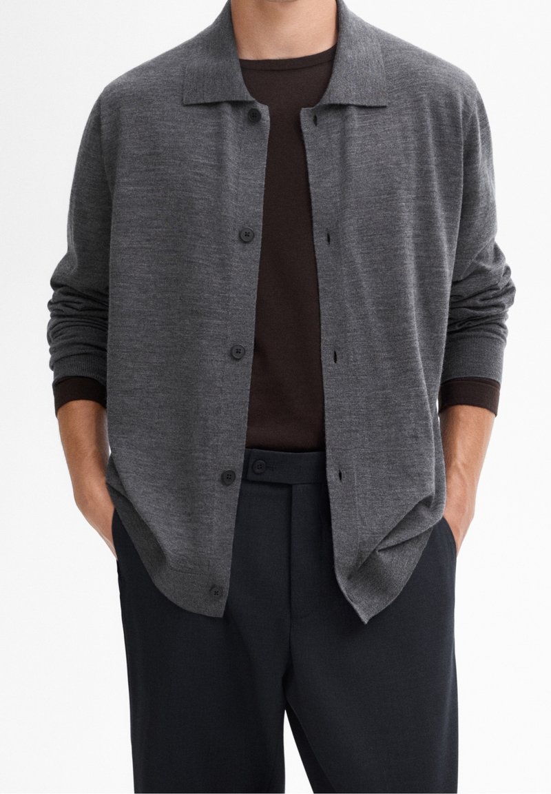 Homme portant un cardigan gris foncé boutonné sur une chemise marron foncé avec un pantalon noir, mains dans les poches, sur fond blanc uni.