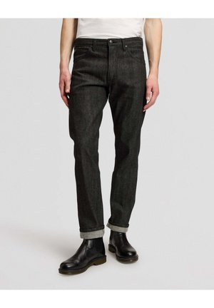 Mann trägt dunkelgraue hochgekrempelte Jeans und schwarze Leder-Stiefeletten, steht vor einem einfarbig hellen Hintergrund.
