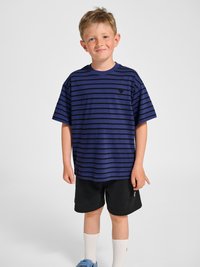 Smilende dreng iført en blå og sort stribet t-shirt, sorte shorts, hvide sokker og blå sneakers, stående foran en ensfarvet baggrund.