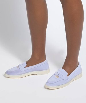 Personne portant des mocassins violets clairs sans lacets avec des semelles blanches, debout sur une surface blanche sur un fond uni.