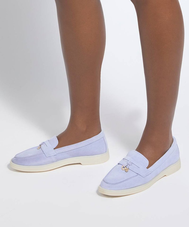 Personne portant des mocassins violets clairs sans lacets avec des semelles blanches, debout sur une surface blanche sur un fond uni.