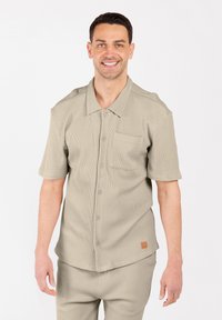 Lachende man in een beige, kortärmige overhemd met knoopsluiting en borstzak, samen met bijpassende broek tegen een witte achtergrond.