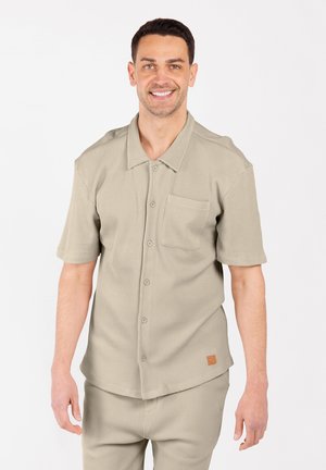 Hombre sonriendo con una camisa de botones de manga corta beige y un bolsillo en el pecho, junto con pantalones a juego, contra un fondo blanco.