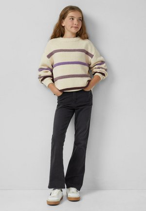 Fleckiger, cremfarbener Pullover mit lila und braunen Streifen, kombiniert mit schwarzen weit geschnittenen Jeans und weißen Sneakern mit braunen Sohlen.