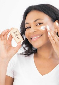 Eine hellpfirsichfarbene Sonnencreme-Flasche mit der Aufschrift "Tägliche Sonnencreme SPF 30", die in einer Hand gehalten wird. Die andere Hand trägt ein cremiges, weißes Produkt auf die Wange auf.