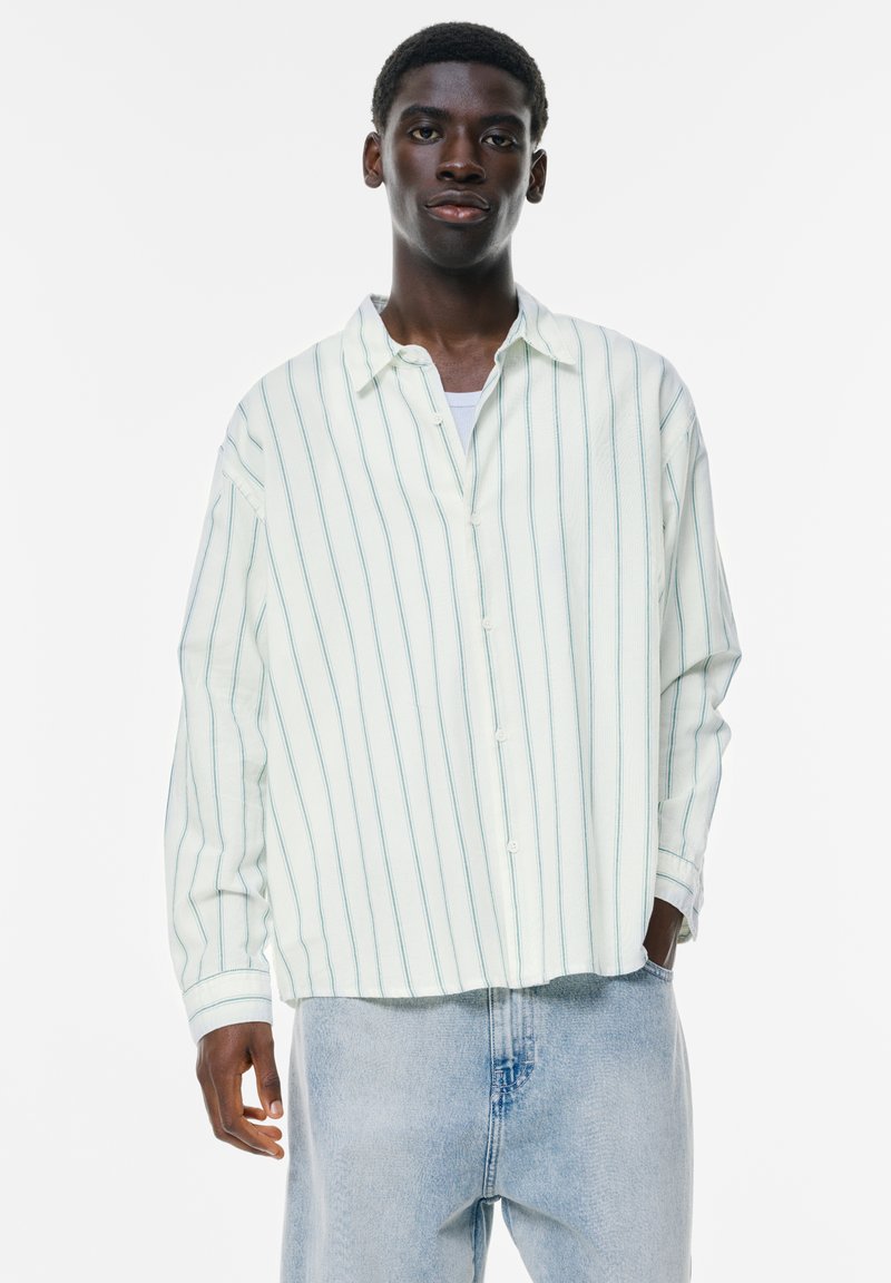Hellgrün gestreiftes Hemd mit entspannter Passform, Knopfleiste und langen Ärmeln. Kombiniert mit hellblauen Jeans.