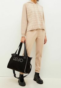 Beige hoodie met logopatroon, beige broek en zwarte, getextureerde shopper met witte accenten en verstelbare banden. Zwarte veterlaarzen.