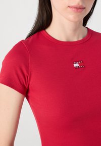 Gros plan d'une personne portant un t-shirt rouge à côtes ajusté, avec un petit logo "Tommy Jeans" sur la poitrine, ayant une peau lisse et des cheveux longs.