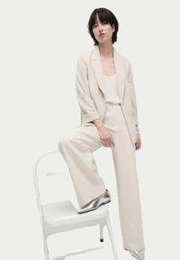 Blazer beige e pantaloni a gamba larga in tessuto leggero; abbinati a una maglia bianca e scarpe argentate; seduto su una sedia bianca.