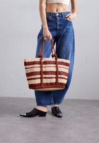 Torba na zakupy z plecionej słomy w czerwono-beżowe paski, z solidnymi brązowymi uchwytami i okrągłym dnem. Modelka ma na sobie luźne dżinsy i czarne klapki.