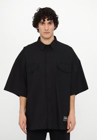 Black short-sleeve button-up shirt made of woven fabric featuring two chest pockets, a curved hem, and a visible brand label on the side.