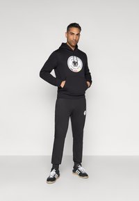 Fekete kapucnis pulóver kör alakú logóval és kenguruzsebbel, hozzá illő, lefelé szűkülő melegítőnadrággal; fekete sportcipőkkel kiegészítve.