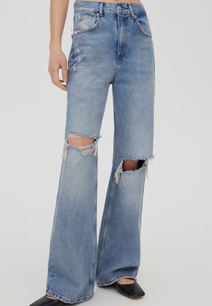 Lichtblauwe jeans met hoge taille en brede pijpen met grote gescheurde gaten op beide knieën, gedragen met zwarte schoenen met vierkante neus op een witte achtergrond.