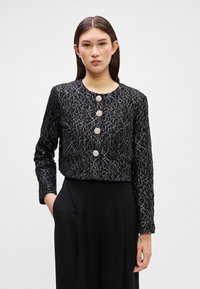 Giacca nera corta con un intricato motivo in pizzo argento, caratterizzata da quattro bottoni rotondi decorativi sul davanti e maniche lunghe.