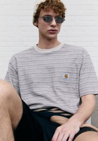 Gray striped short-sleeve t-shirt with a chest pocket and logo patch. Worn with black shorts, exposing skin. Light background.