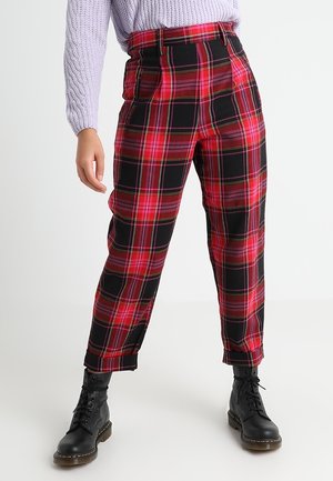 Pantalons écossais rouges et noirs avec une taille haute, des jambes fuselées et des plis à l'avant. Portés avec des bottes noires à lacets et un pull léger couleur mauve.