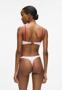 Tamsiaodės odos ir plaukų į kuodą surištas moteris, vilkinti baltą ir raudonų širdelių rašto liemenėlę ir stringus, stovi atsisukusi nugara priešais baltą foną.