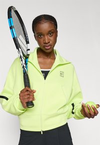 Veste zippée vert fluo avec des accents noirs, portée sur un haut noir. Tenant une raquette de tennis et une balle de tennis verte.