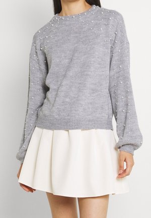 Femme portant un pull gris clair orné de perles et une jupe plissée blanche, montrée des épaules jusqu'à mi-cuisse.