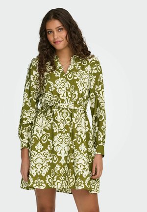 Vrouw met krullend bruin haar die een groen-witte, lange mouwen jurk met patroon en een strik in de taille draagt, staand tegen een neutrale achtergrond.