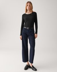 Frau trägt ein schwarzes, langärmeliges Rippenshirt, dunkelblauen weiten Jeans mit Gürtel und schwarze Ledermokassins, stehend vor einem hellen Hintergrund.