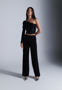 Top court en sequins noirs à une manche, associé à un pantalon noir ample et des chaussures à bout pointu métalliques sur un fond neutre.