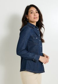 Camisa de mezclilla azul oscuro con mangas largas, cierre de botones en la parte frontal y dos bolsillos en el pecho. Presenta un cuello clásico y una costura de contraste sutil.