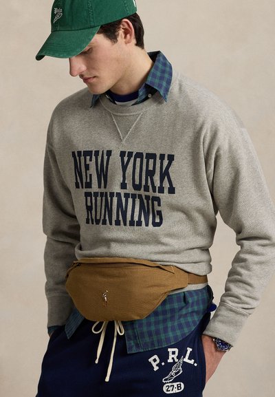 Man draagt een groene pet, grijze trui met de tekst "New York Running", een blauw geruit overhemd, marineblauwe joggingbroek met logo, en een bruine heuptas.