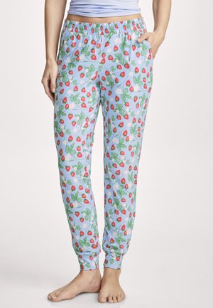 Woman wearing light blue jogger pants with red strawberry and green leaf pattern, hands in pockets, barefoot.