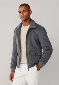 Chaqueta gris de mezcla de lana con cuello de solapa, cremallera frontal y dos bolsillos laterales. Estilizada sobre un suéter de punto color crema, combinada con pantalones blancos.