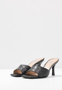 Sandalias de tacón negro con parte superior de cuero trenzado, diseño abierto en la parte delantera, puntera cuadrada y tacón delgado. Acabado suave con etiqueta beige en la plantilla.