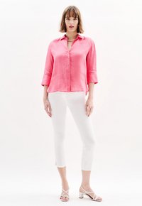 Roze linnen overhemd met knopen en driekwartmouwen, gecombineerd met witte cropped broeken. Model draagt witte sandalen met hakken en bandjes.
