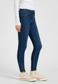 Jean skinny en denim bleu foncé avec une texture lisse, présentant une fermeture éclair au niveau de la cheville et un design classique à cinq poches.