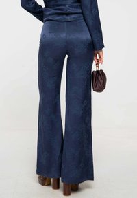 Pantalons bleu marine à flare texturés avec un motif subtil. Associés à un sac à main rond marron foncé et des chaussures à talons blocs marron.
