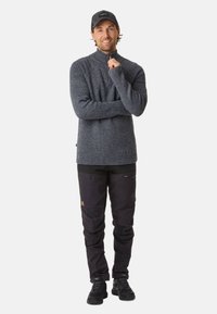 Un pull col roulé gris à côtes associé à un pantalon cargo noir avec des poches zippées et une étiquette marron, complété par des baskets noires et une casquette.