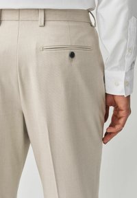 Gros plan de la poche d’un pantalon beige avec détail de bouton noir et une main à côté de la hanche, porté avec une chemise blanche à manches longues.