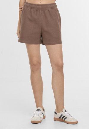 Persoon draagt bruine gestructureerde korte broek met elastische taille en witte Adidas sneakers met zwarte strepen, staand op een witte achtergrond.